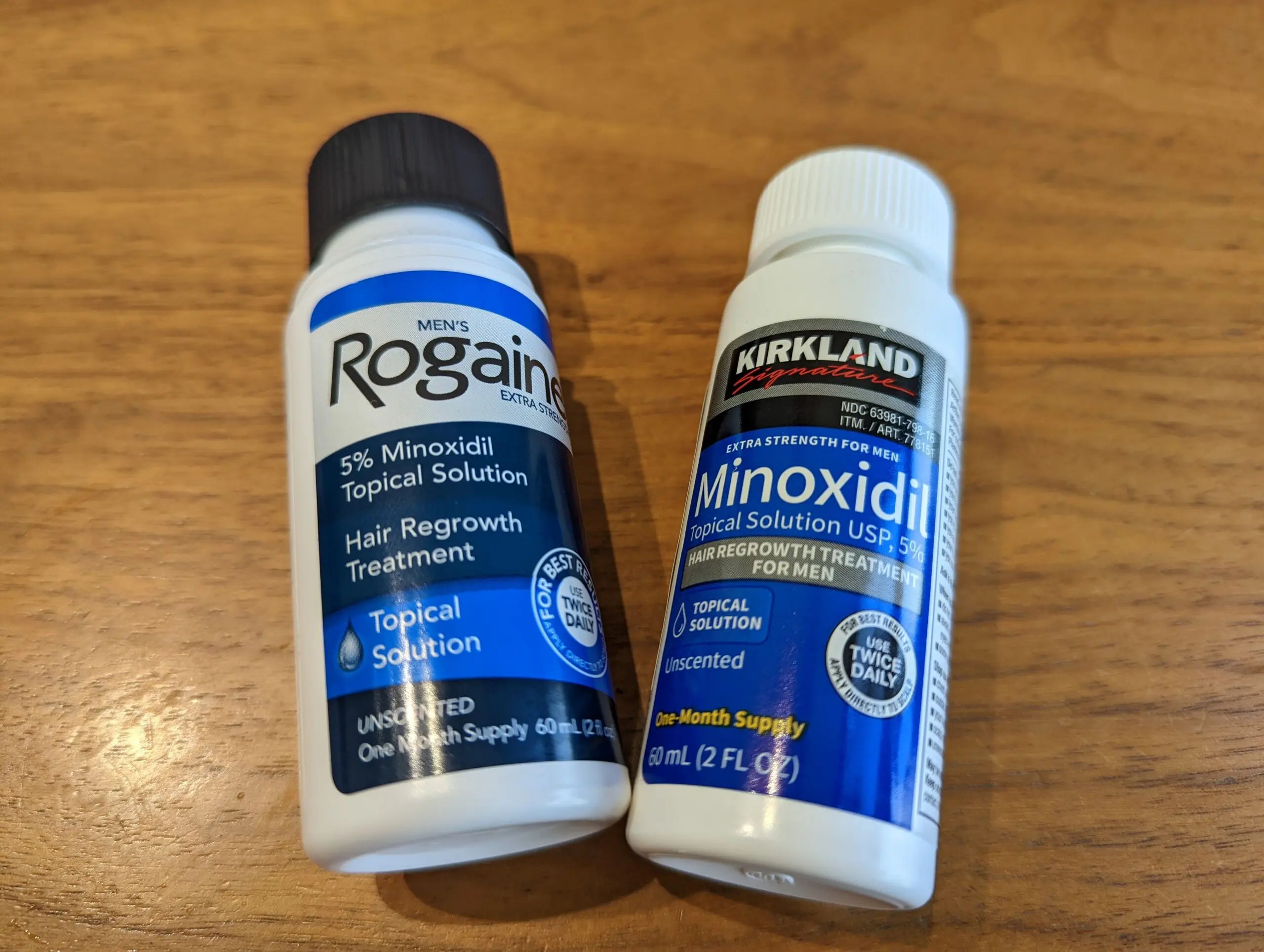 Kirkland Signature Minoxidil 5% 與 Men's Rogaine 落建生髮液實體並排比較圖：右側為好市多自有品牌 Kirkland，左側為原廠落建，兩者放置於乾淨木紋桌面上展示外觀差異。